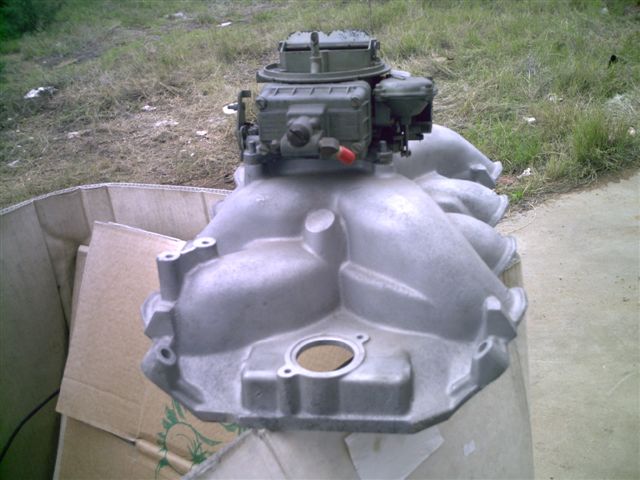 Aluminum intake manifold with a Holley carburetor, resting in a cardboard box outdoors