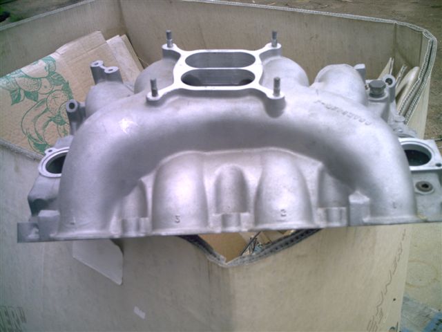 Aluminum intake manifold with dual carburetor openings, sitting in a cardboard box