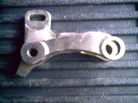 1964 Ford 427 - A metal linkage part is shown against a textured background. The part has two circular openings and a curved arm.