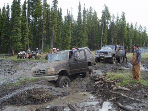 1995 Ford Bronco - Several off-road vehicles are stuck in a muddy area with trees in the background.