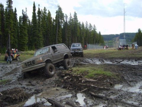 1995 Ford Bronco - A muddy Ford Bronco tackles an off-road course, with a second truck visible in the distance and spectators nearby.