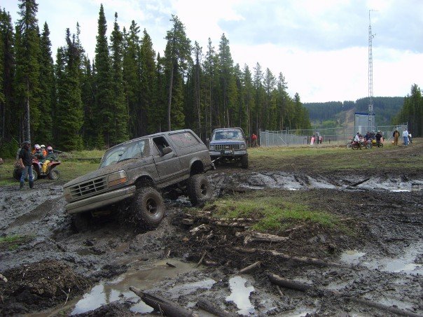 Mud-covered Ford Bronco navigating a muddy track, with another truck behind it and trees in the background