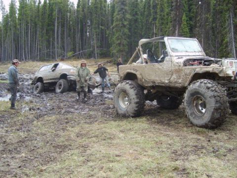1995 Ford Bronco - Two off-road vehicles are stuck in the mud with people around them in a wooded area.