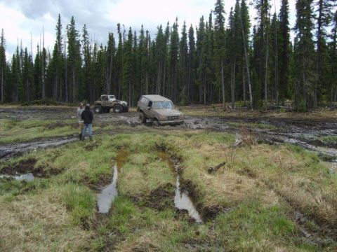 1995 Ford Bronco - A muddy off-road scene with a Ford Bronco stuck in the mire, accompanied by another vehicle and onlookers.