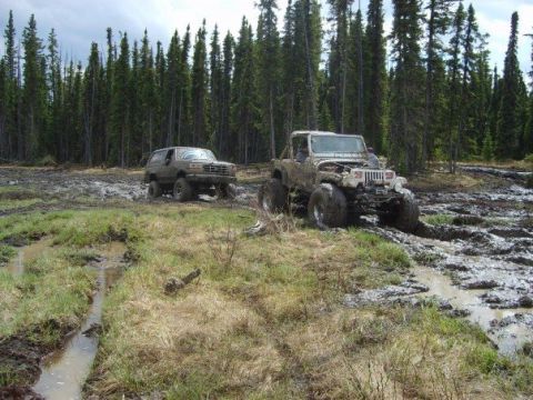 1995 Ford Bronco - Two off-road vehicles navigate a muddy trail through a dense forest.