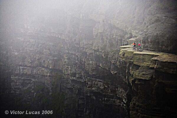 Two people on mountain bikes ride along a narrow cliff edge with a foggy background