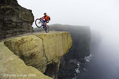 1968 Ford Mustang - A mountain biker balances on a cliff edge, with the ocean below and fog obscuring the background.