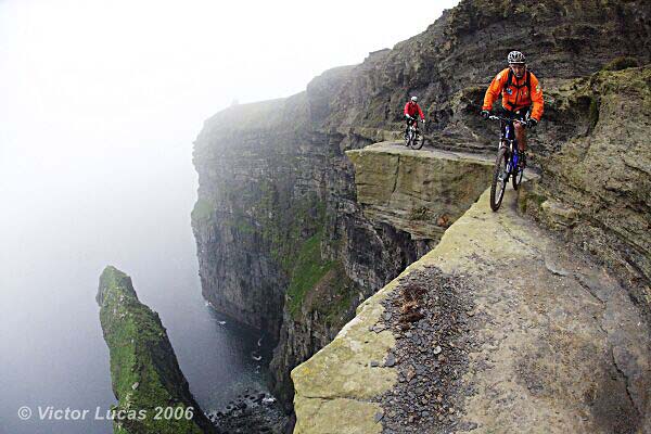 Two mountain bikers ride along a narrow cliff edge overlooking the ocean