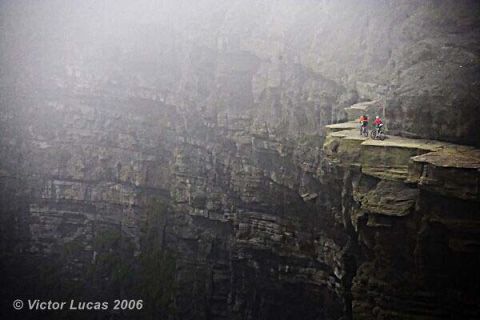 1968 Ford Mustang - Cyclists navigate a precarious cliff edge, with fog obscuring the vast landscape.