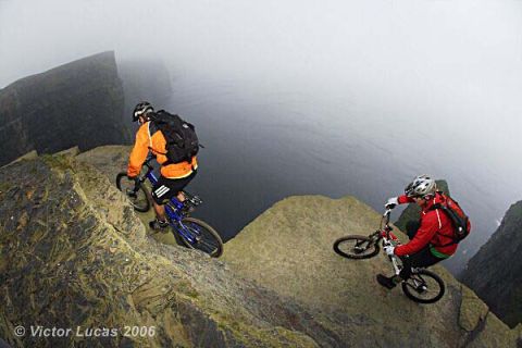 1968 Ford Mustang - Two cyclists navigate a treacherous cliffside path, with a vast expanse of water below and fog obscuring the horizon.