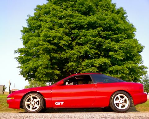 1993 Ford Probe - A red coupe with a GT badge is parked on a gravel surface with a large green tree in the background.