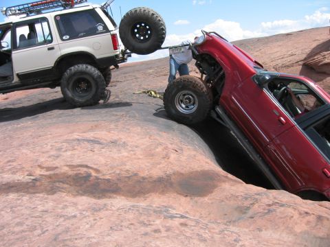 1997 Ford Ranger - A red off-road vehicle is precariously stuck in a rock crevice, with a white SUV attempting to pull it out.