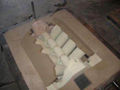 1972 Ford Ranchero - Open foundry sand-casting mold in a steel flask on a shop table, with tan molding sand and a row of white cylindrical cores set in the cavi…