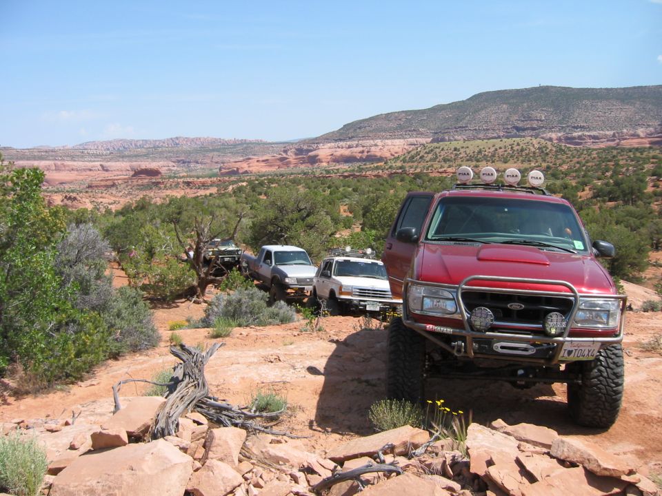 Red Ford Explorer with off-road modifications leads a convoy of trucks on a desert trail