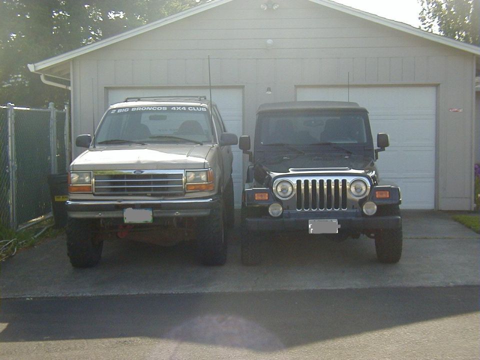 Tan Ford Explorer and black Jeep Wrangler parked side-by-side in front of a two-car garage. Explorer has a "2 BIG BRONCOS 4X4 CLUB" sticker on windshield