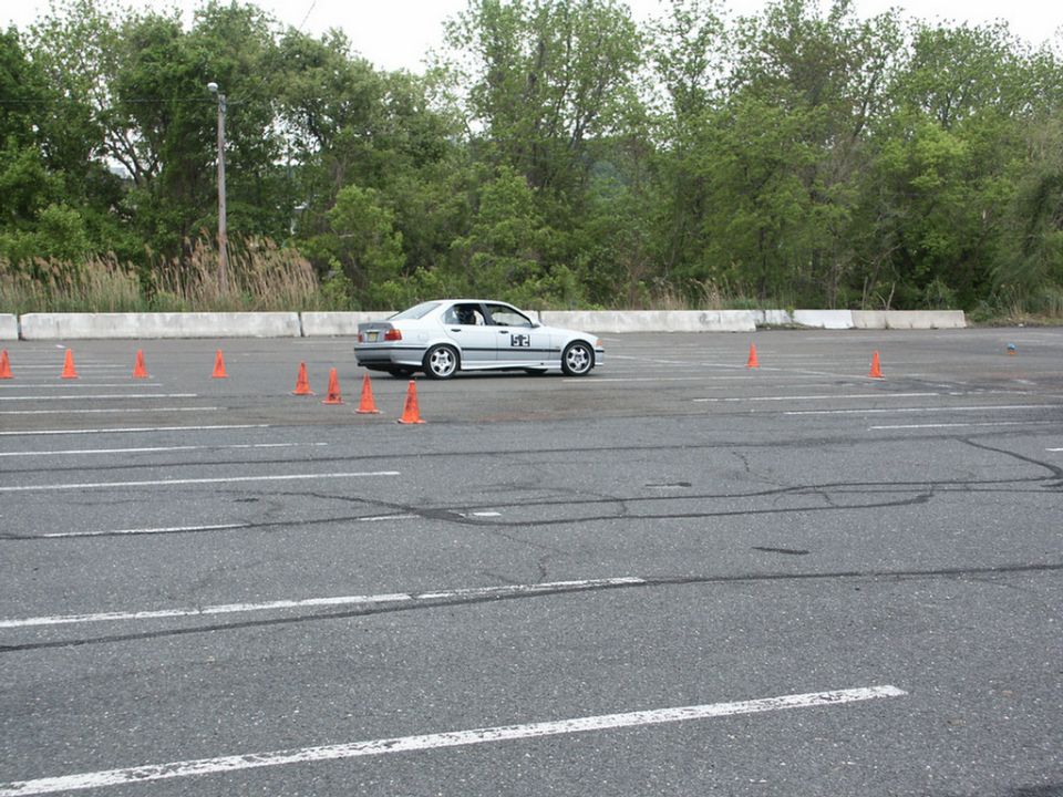 Silver BMW E36 sedan with racing numbers on the door, navigating an autocross course marked by orange cones