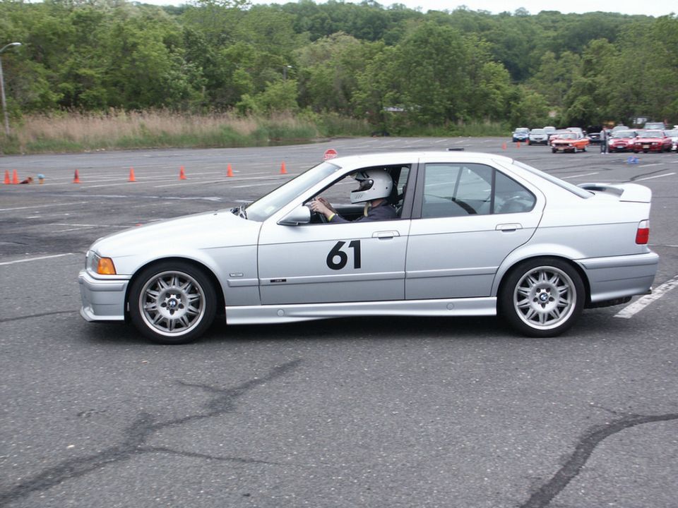 Silver BMW E36 sedan, number 61 on door, driver wearing helmet. Autocross course with cones in background