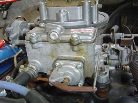 1979 Ford Bronco - Close-up of a cast-aluminum two-barrel carburetor still mounted on the intake in an engine bay, showing the fuel-bowl side with accelerator…