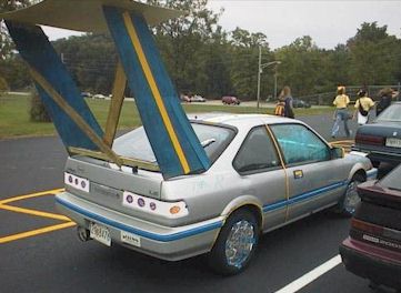 1995 Ford Bronco - Low-resolution three-quarter rear shot of a silver two-door compact hatchback in a parking lot, fitted with an enormous multi-tier rear win…