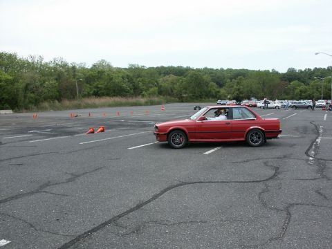 2001 Mercury Grand Marquis - A red BMW E30 coupe navigates a course marked by orange cones in a parking lot, with other vehicles visible in the distance.