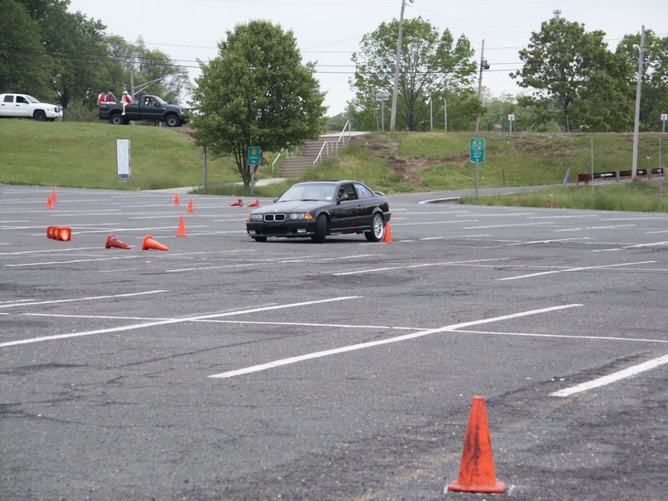 Black BMW coupe driving through an autocross course marked by orange cones. Another truck and SUV are visible on a grassy incline in the background