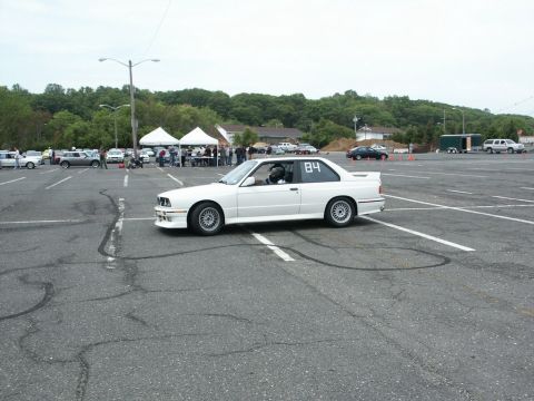 2001 Mercury Grand Marquis - A white BMW E30 coupe with racing number 84 is seen at an event with other vehicles and tents in the background.