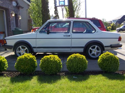 1984 Volkswagen Jetta - A white coupe with distinctive black side trim is parked in front of a garage and manicured bushes.