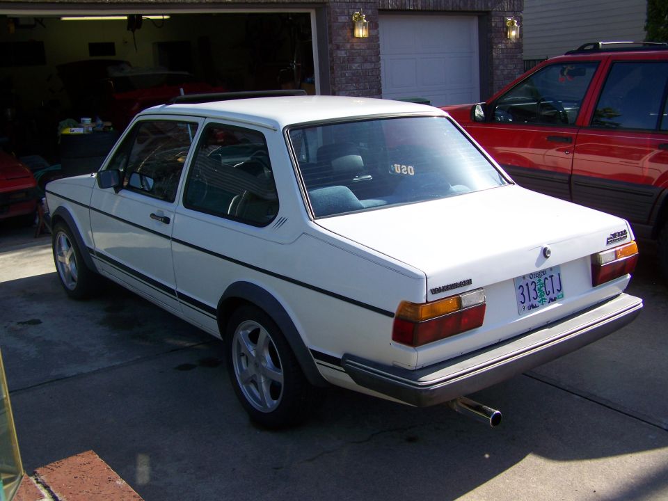 White 1984 Volkswagen Jetta parked in a driveway, rear 3/4 view. A red SUV is parked to the right