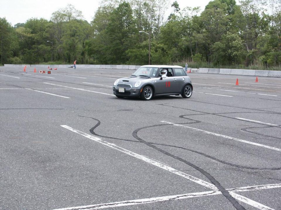 Gray Mini Cooper S with a white roof and red racing number on the door, driving on an asphalt track with tire marks
