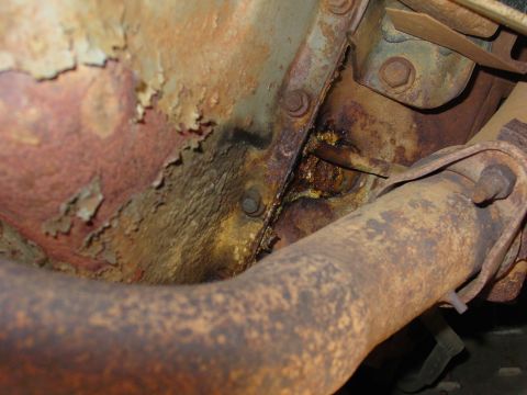1994 Ford Bronco - Tight underside detail of a heavily rusted vehicle floor and frame area, with flaking paint, scale rust, and a corroded exhaust pipe curvin…