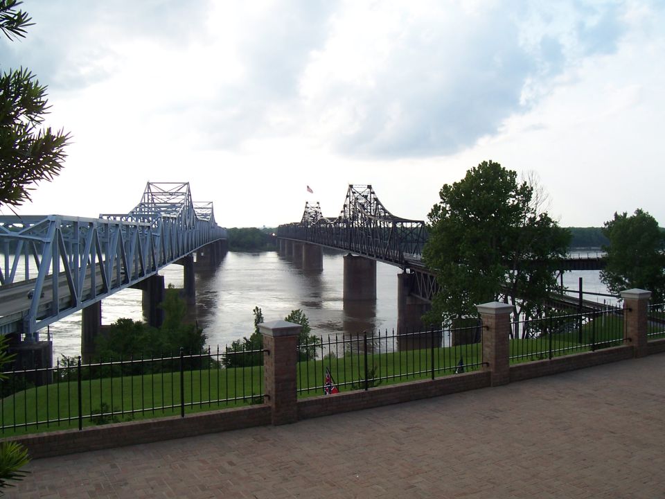 Two bridges cross a wide river under a cloudy sky. One bridge is light blue, the other is dark
