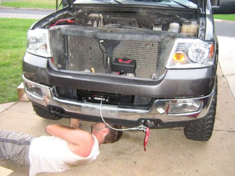 1986 Ford Bronco - A person works on the front of a gray Ford F-150, which has a winch mounted below the bumper.