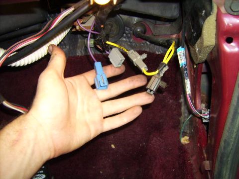 1982 Ford LTD Crown Victoria - A hand holds up several unplugged wiring connectors in a car's driver footwell, a blue plastic plug, white and gray square connectors on ye…