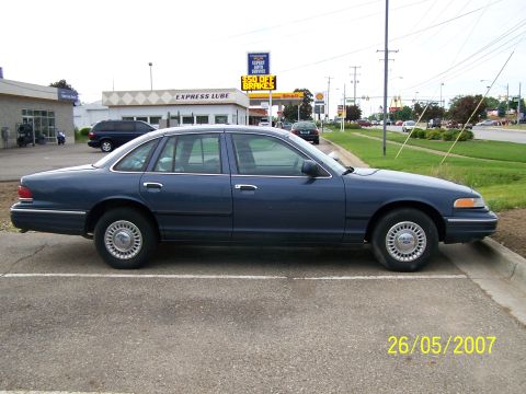 2003 Ford Crown Victoria - A blue 2003 Ford Crown Victoria is parked in a lot with a business and street in the background.