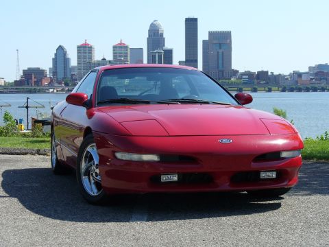 1991 Ford Probe - A red 1993 Ford Probe is parked near a body of water, with a city skyline visible behind it.