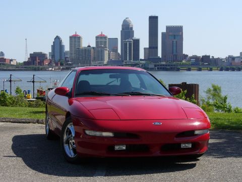 1991 Ford Probe - A red 1991 Ford Probe is parked by the water with a city skyline in the background.