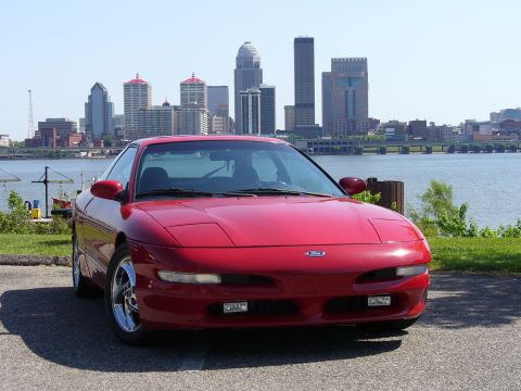 1991 Ford Probe - A red 1991 Ford Probe is parked near a body of water with a city skyline visible behind it.
