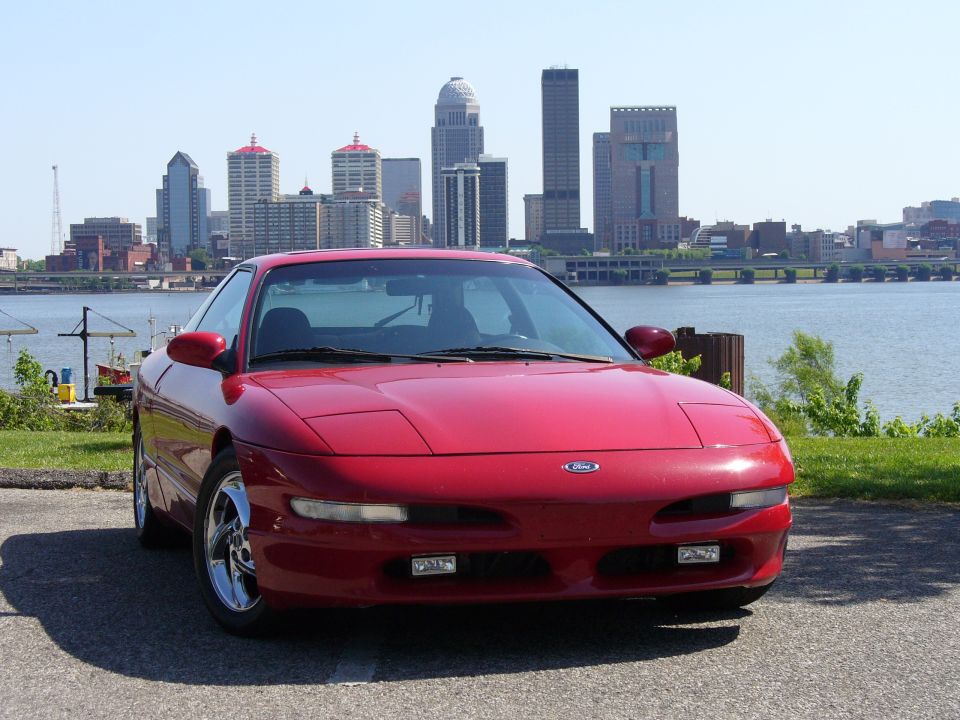 Red 1991 Ford Probe parked by a river with a city skyline in the background