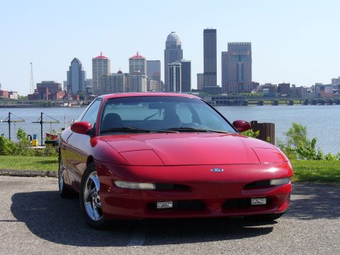 1991 Ford Probe - A red Ford Probe is parked near a body of water with a city skyline in the distance.