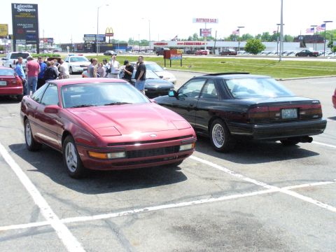1991 Ford Probe - Two coupes, a red Ford Probe and a black Mazda MX-6, are parked side-by-side in a lot with other vehicles and buildings in the background.