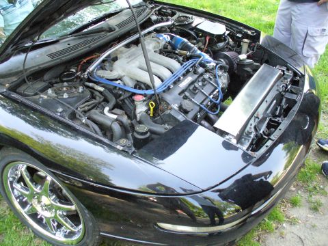 1991 Ford Probe - The engine bay of a black car, possibly a Mazda MX-6, is open for inspection, revealing its V6 engine and custom modifications.