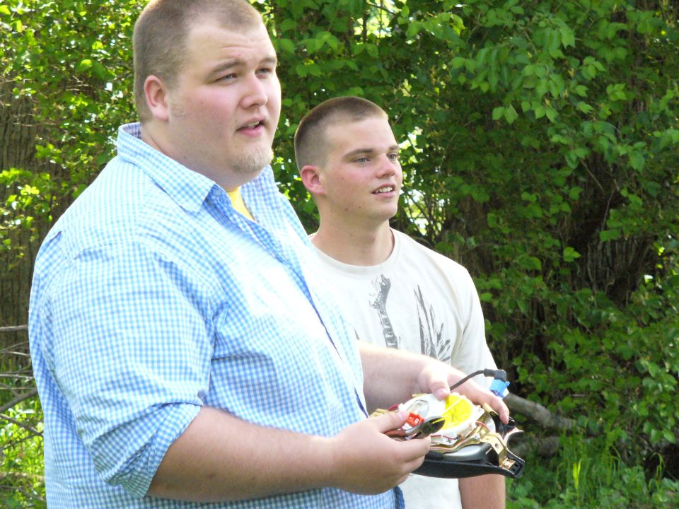 Two men stand outdoors; one holds a disassembled remote control car part
