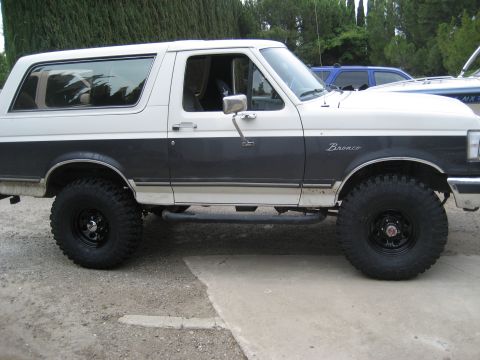 1989 Ford Bronco - Passenger-side profile of a two-tone Ford Bronco, white over dark gray-brown with chrome wheel-arch trim and Bronco script on the fender. I…