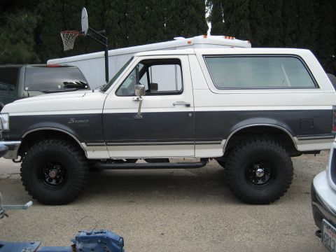 1989 Ford Bronco - Driver-side profile of a lifted white-over-gray Ford Bronco with the white removable hardtop in place, Bronco fender script, chrome west-co…