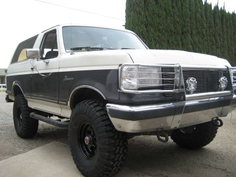 1989 Ford Bronco - Close three-quarter front view of a lifted white-over-dark-blue Ford Bronco with hardtop, parked on concrete beside a tall cypress hedge. I…