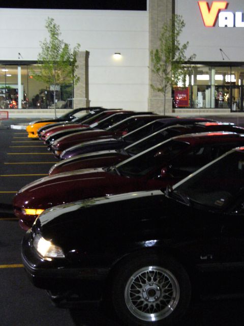 1991 Ford Probe - A line of cars, including a black 1991 Ford Probe, are parked in a lot at night.