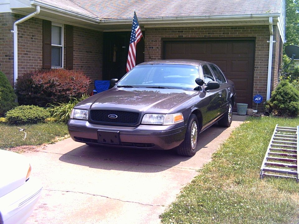 Gray Ford Crown Victoria sedan parked in driveway of brick house with American flag