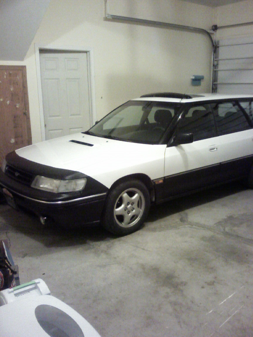 White Subaru Legacy wagon with black hood bra parked in a garage, front three-quarter view