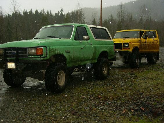 Lifted green Ford Bronco with white top beside a yellow Bronco in falling snow
