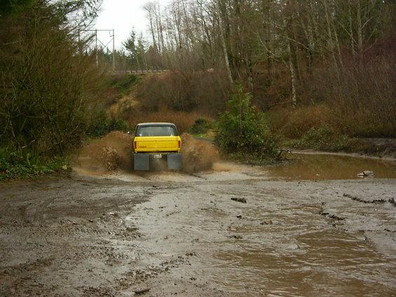 Yellow lifted Ford truck spraying mud through a flooded pit, rear view